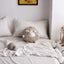 Khaki round sheep plush toy on a white bed with pom-pom pillows and a small sheep companion.