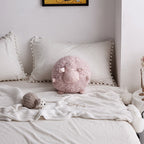 Pink round sheep plush toy on a white bed with decorative pillows and a small grey sheep accent.