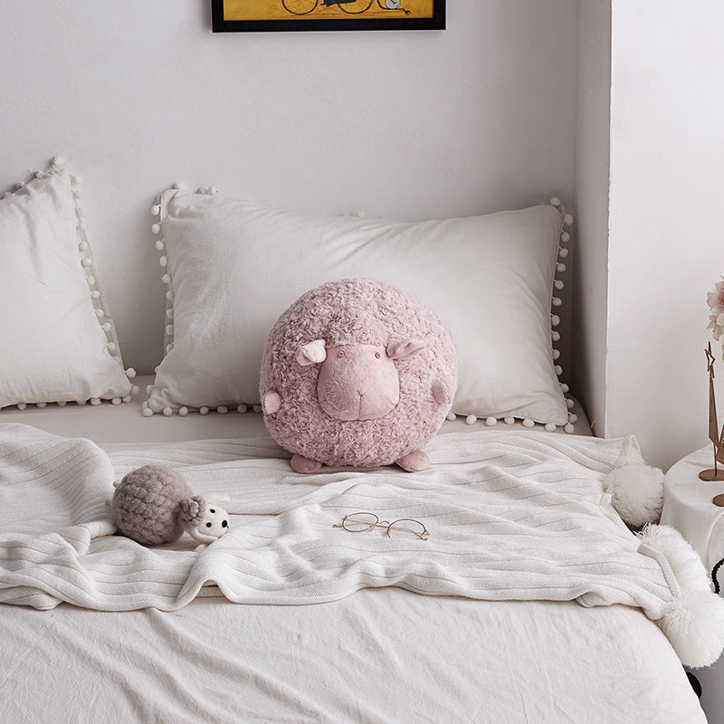 Pink round sheep plush toy on a white bed with decorative pillows and a small grey sheep accent.