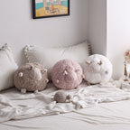 Three round sheep plush toys in brown, pink, and white displayed on a cozy bed with pillows.