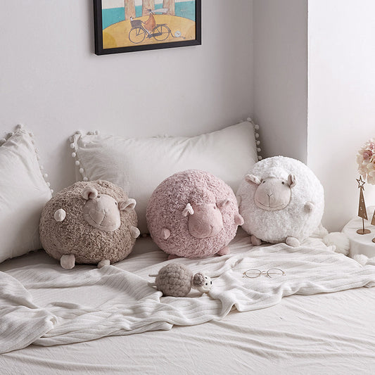 Three round sheep plush toys in brown, pink, and white displayed on a cozy bed with pillows.