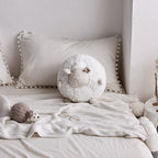 White round sheep plush toy on a cozy bed with tassel pillows and a small grey sheep accent.