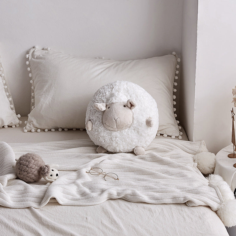 White round sheep plush toy on a cozy bed with tassel pillows and a small grey sheep accent.