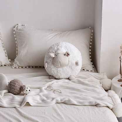 White round sheep plush toy on a cozy bed with tassel pillows and a small grey sheep accent.